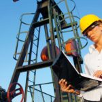 an oil refinery worker checks her laptop computer in front of a backdrop of oil refinery infrastructure. The woman has a serious look on her face.