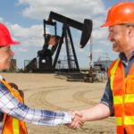 Two workers shake hands in front of an oil rig on the successful completion of a deal.