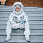 a man in full astronaut suit sits forlornly on a set of concrete steps with a sorrowful look on his face beneath his rounded space helmet.