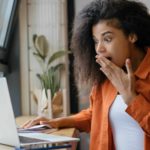 A young woman holds her hand to her mouth in surprise as she reads something on her laptop.