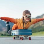 Kid with arm spread out on a luggage bag, riding a skateboard.