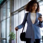 A corporate-looking woman looks at her mobile phone as she pulls along her suitcase in another hand while walking through an airport terminal with high glass panelled walls.