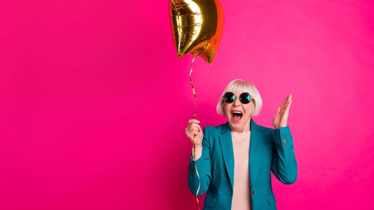 A cool older woman wearing sunglasses celebrates at her party with a gold balloon.