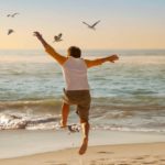 A boy leaps and flaps his arms as he tries to fly with some birds on the shoreline of the beach.