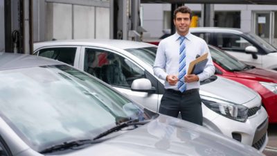 a car dealer stands amid a selection of cars parked in a showroom while he is holding a set of keys and paperwork in his other hand.