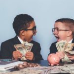 two young boys dressed in business suits and wearing spectacles look at each other in rapture with wide open mouths and holding large fans of banknotes with other banknotes, coins and a piggybank on the table in front of them and a bag of cash at the side.