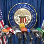 a picture of the US federal reserve podium for making media announcements complete with US flag and federal reserve flag in the background and a large array of microphones set up.