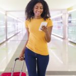 A smiling woman looks at her phone as she walks with her suitcase inside an airport.