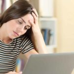 a woman looks exhausted and overwhelmed as she slumps forward into her hand while looking at her laptop screen.