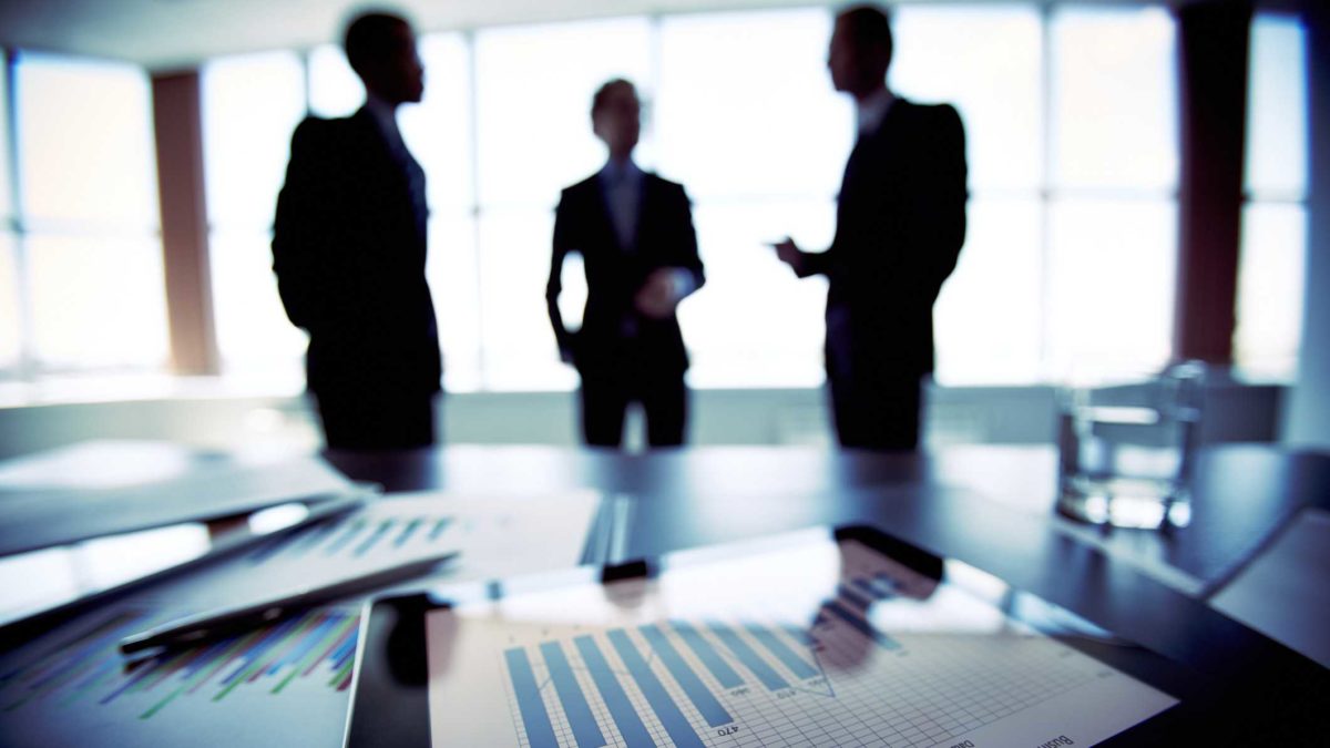 three businessmen stand in silhouette against a window of an office with papers displaying graphs and office documents on a desk in the foreground.