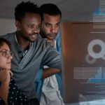 Three people gather around a large computer screen where they are looking at something that is captivating their interest with a graphic image of data and digital technology material superimposed to the right hand third of the image.