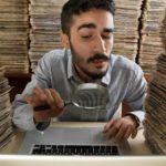 a man surrounded by huge piles of paper looks through a magnifying glass at his computer screen.