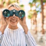 A woman standing on the street looks through binoculars.