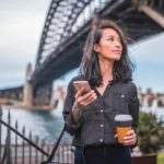 A woman wearing dark clothing and sporting a few tattoos and piercings holds a phone and a takeaway coffee cup as she strolls under the Sydney Harbour Bridge which looms in the background.