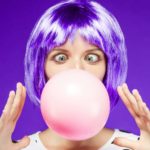 a woman with bright artificially coloured hair blows a large bubble gum bubble from her mouth with her eyes wide open and holding her hands either side of it.
