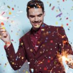 a man holds a firework sparkler in both hands as a shower of sparkly confetti falls from the sky around him as he smiles and closes his eyes in a celebratory scene.