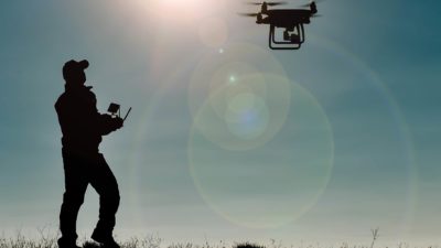 A silhouette shot of a man holding a control in his hands and watching as a drone hovers overhead with sunrays coming from the sky.