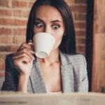 A woman looks shocked as she drinks a coffee while reading the paper.