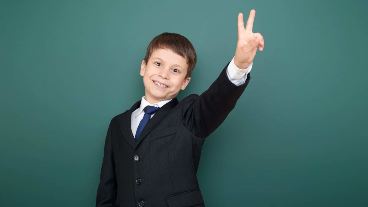 a young boy dressed up in a business suit and tie has a cute grin and holds two fingers up.