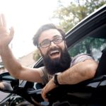 A man leans out of his car window with a massive smile on his face and waves.