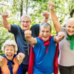 A group of older people wearing super hero capes hold their fists in the air, about to take off.