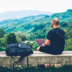 A man sits on a bench atop a mountain with a laptop, making investments with a green ESG mind.