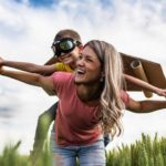 A little boy in flying goggles and wings rides high on his mum's back with blue skies above.