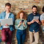 A group of young people lined up on a wall are happy looking at their laptops and devices as they invest in the latest trendy stock.