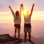 Two people climb to the summit and raise their arms in success as the sun rises brightly over the mountains.