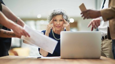 A mature businesswoman has people demanding things from her from all angles, and she looks stressed.