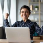 A woman gives two fist pumps with a big smile as she learns of her windfall, sitting at her desk.