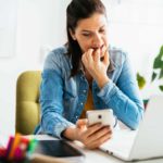 A worried woman looks at her phone and laptop, seeking ways to tighten her belt against inflation.