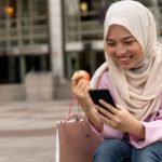 A young woman wearing an Islamic tradition headscarf and jeans sits in an urban environment with an apple in one hand and her phone in the other with a smile on her face.