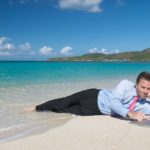 A man in business pants, a shirt and a tie lies in the shallows of a beautiful beach as he consults his laptop on the shore, just out of the water's reach.