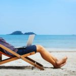 An ASX dividend investor lies back in a deck chair with his hands behind his head on a quiet and beautiful beach with blue sky and water in the background.