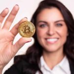 A woman holds a bitcoin token in her hand as she smiles at the camera in the background.