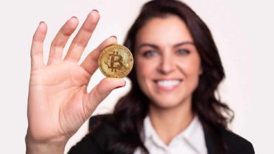 A woman holds a bitcoin token in her hand as she smiles at the camera in the background.