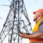 A male electricity worker in hard hat and high visibility vest stands underneath large electricity generation towers as he holds a laptop computer and gazes up at the high voltage wires overhead.