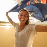 A fresh-faced young woman holds an Australian flag aloft above her head as she smiles widely on a beach as though celebrating a national day or event where Australia has been successful.