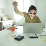 A man with a beard and wearing dark sunglasses and a beanie head covering raises a fist in happy celebration as he sits at is computer in a home environment.