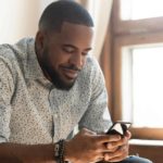 A man leans forward over his phone in his hands with a satisfied smirk on his face although he has just learned something pleasing or received some satisfying news.