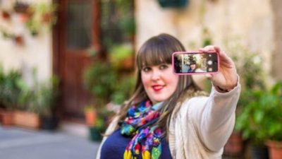 A woman in colourful outfit holds up a phone to take a selfie.