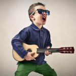 A young kid with dark glasses rocks out with a guitar.