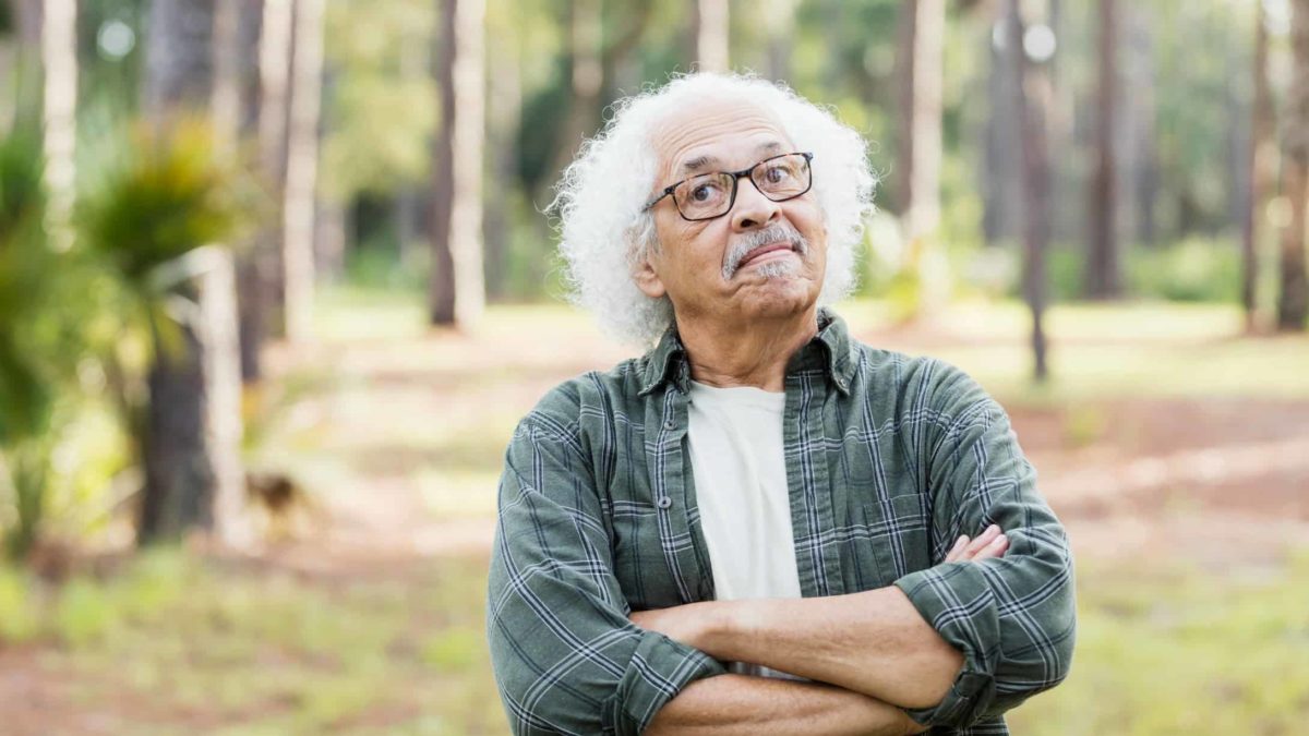 Un vecchio con i capelli bianchi e ondulati incrocia le braccia in un gesto testardo mentre si trova con aria di sfida in un ambiente all'aperto.