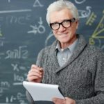 A grey-haired mature-aged man with glasses stands in front of a blackboard filled with mathematical workings as he holds a pad of paper in one hand and a pen in the other and stands smiling at the camera.