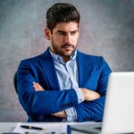 An unhappy man in a suit sits at his desk with his arms crossed staring at his laptop screen as the PointsBet share price falls