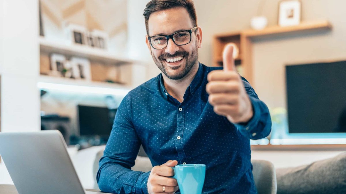 A man holding a cup of coffee gives a thumbs up and smiles while working on a laptop.