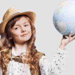 A cute young girl wears a straw hat and has a backpack strapped on her back as she holds a globe in her hand with a cheeky smile on her face.