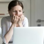 A woman sits at her computer with her hands clutched her the bottom of her face as though she may be biting her fingermails with a worried expression in her eyes and frown lines visible.