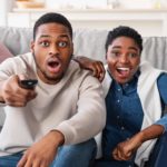 A couple stares at the tv in shock, one holding the remote up ready to press.